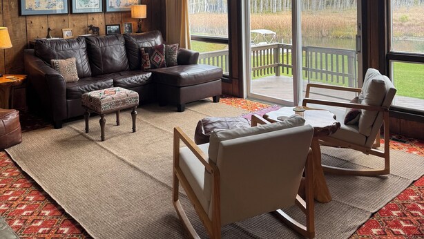 Una sala de estar acogedora con un sofá de cuero, una mesa de centro de madera y un tapete estampado. Amplias ventanas ofrecen vistas a una terraza y árboles.
