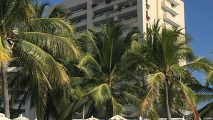 Een terras aan het strand met witte ligbedden en parasols, palmbomen en een gebouw op de achtergrond.