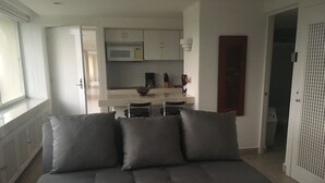 Een moderne woonkamer met een grijze bank, een eethoek met een witte tafel en stoelen, en een keuken met witte kasten en apparatuur.