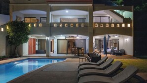 Una casa de dos pisos con piscina, mobiliario exterior e interiores bien iluminados.