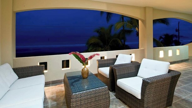 Un balcón con muebles de mimbre, un jarrón con flores y vistas al océano al atardecer.
