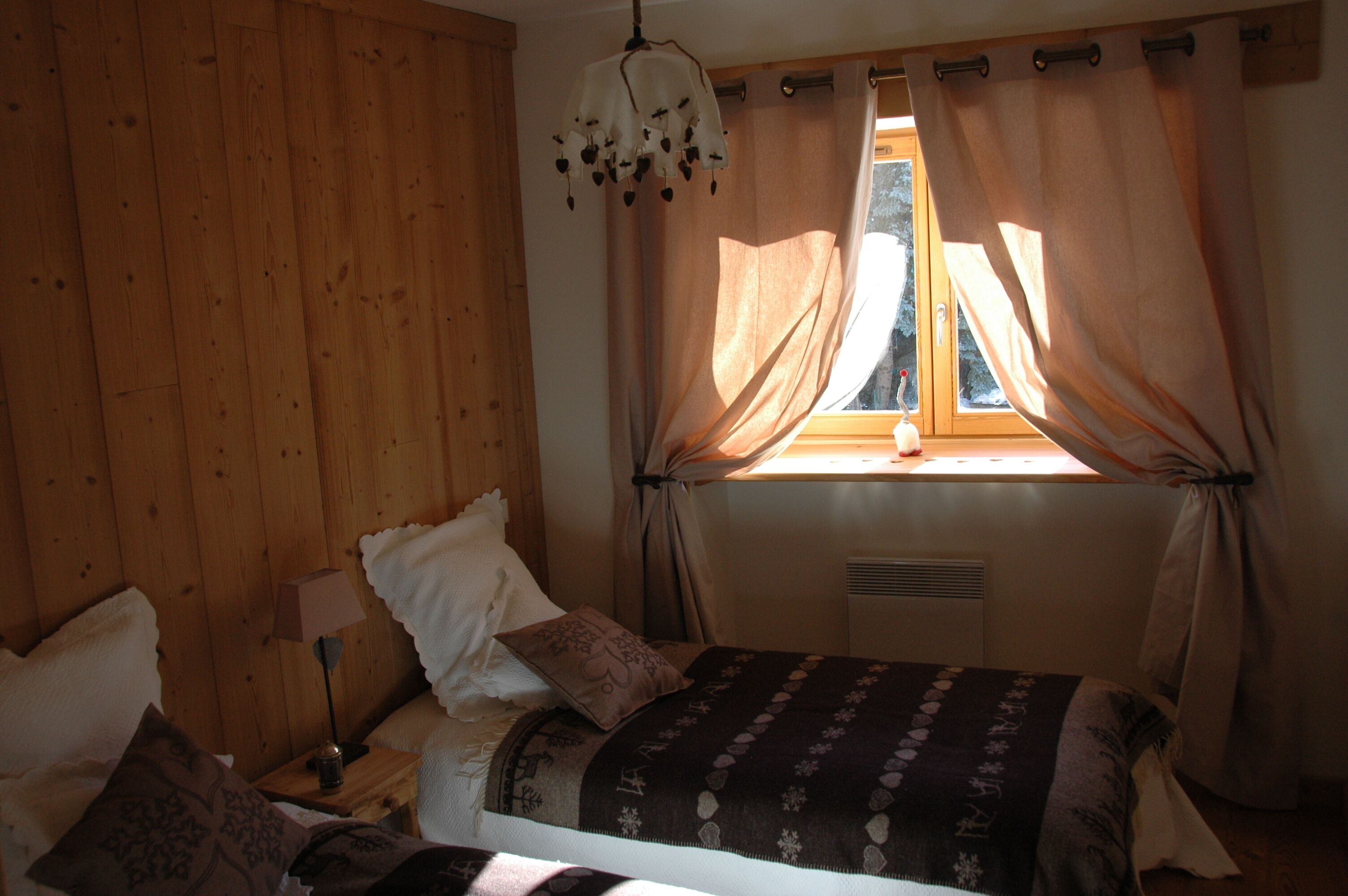A bedroom with a wooden paneled wall, a bed with a patterned bedspread, a bedside lamp, and a window with curtains.
