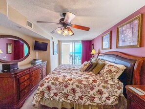 Ein Hotelzimmer mit Bett, Deckenventilator, Kommode, Fernseher und Blick auf das Meer.