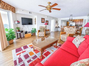 A living room with a red sofa, a coffee table, and a television. There are plants, a ceiling fan, and a kitchen in the background.