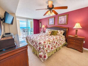 A bedroom with a bed, a desk, a television, a ceiling fan, and a view of the ocean.