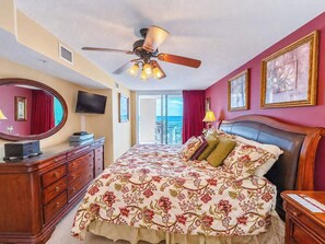Una habitación de hotel con cama, ventilador de techo, televisión, cómoda y balcón con vistas.