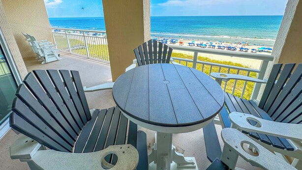 A balcony with a round table and two Adirondack chairs, overlooking a beach with umbrellas and people.