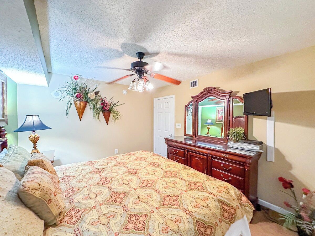 A bedroom with a bed, dresser, ceiling fan, and a television mounted on the wall.