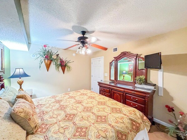 A bedroom with a bed, dresser, ceiling fan, and a television mounted on the wall.