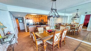 A spacious dining area with a wooden table and chairs, a kitchen in the background, and a chandelier above.