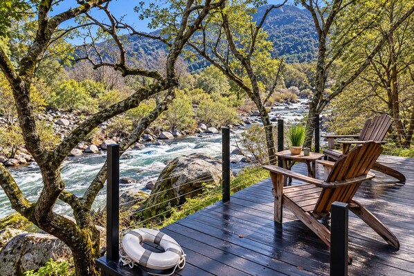 Une terrasse dotée de chaises et d’une table en bois, offrant une vue sur une rivière et un paysage montagneux en arrière-plan.