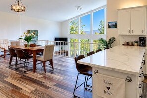 A modern kitchen with a marble countertop, wooden flooring, and a view of greenery through large windows.