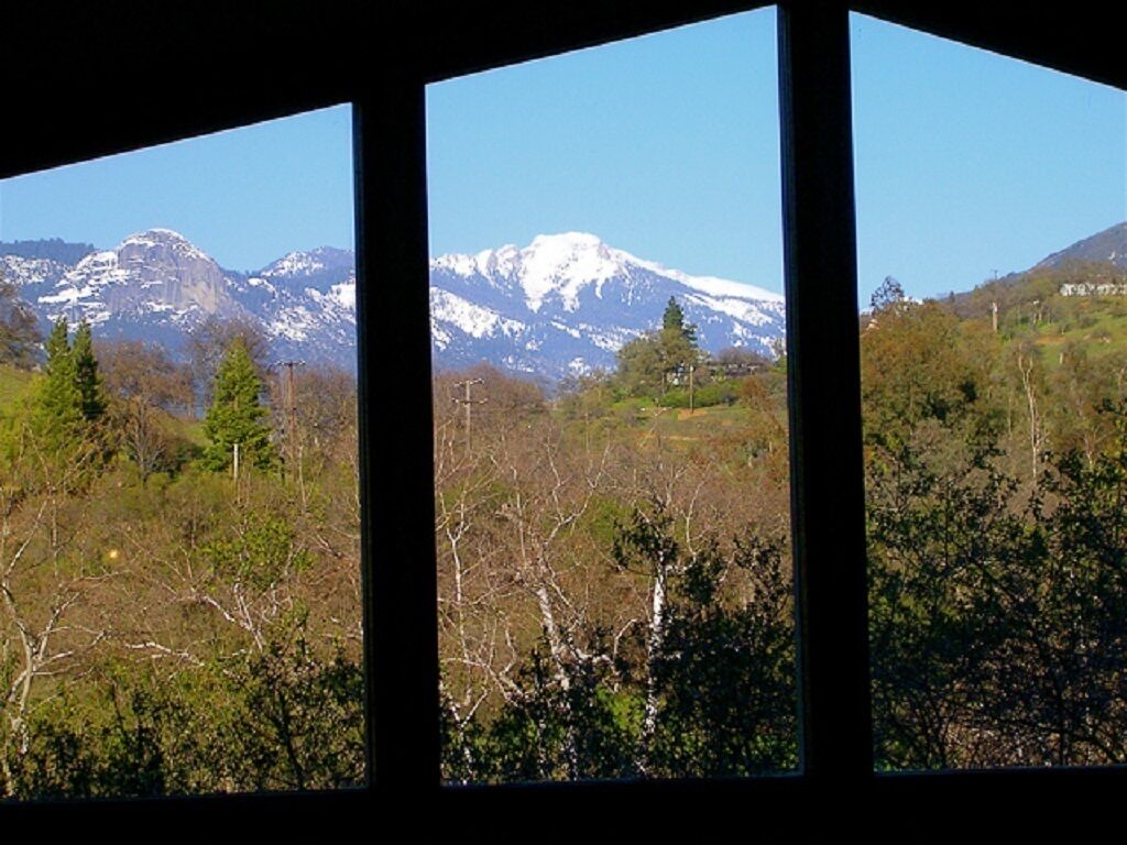 A view through a window with a mountain landscape, including snow-capped peaks and a forested area.