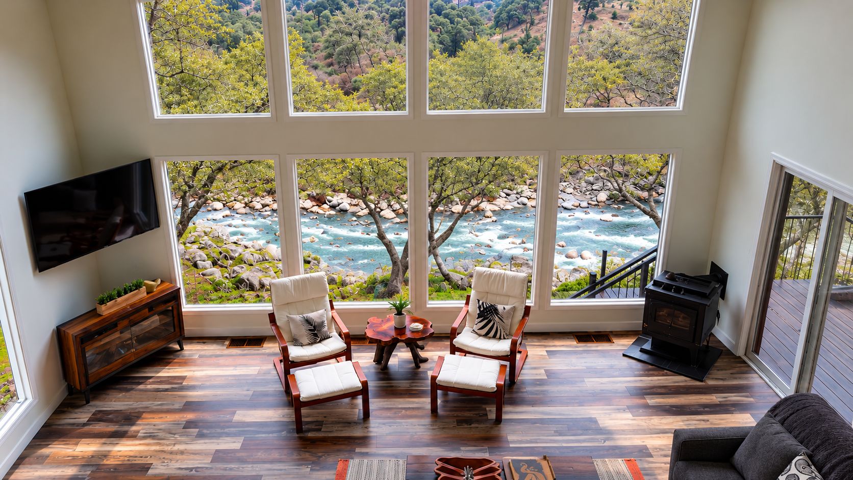 A spacious room with a large window overlooking a river and forest, two armchairs, a TV, and a fireplace.