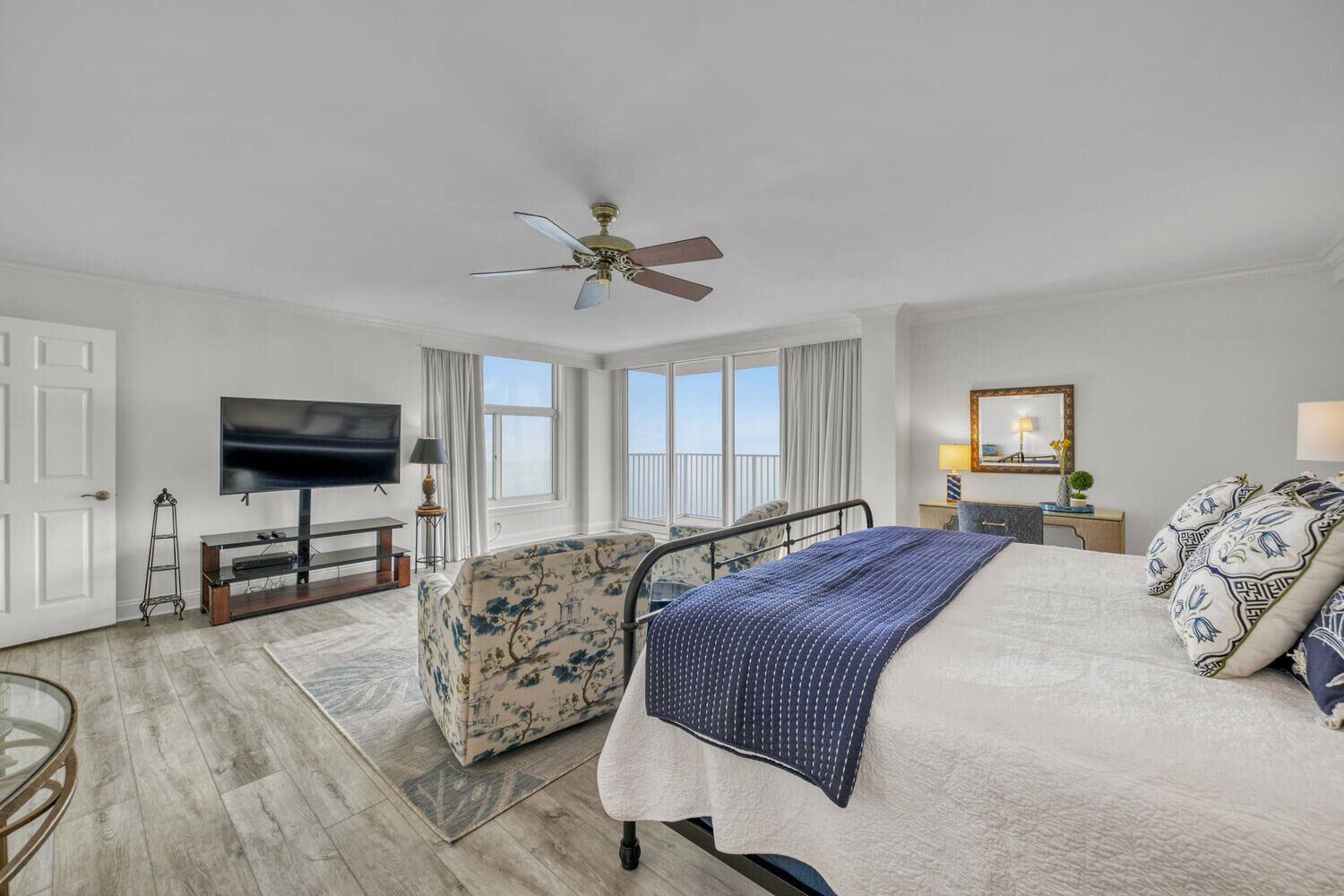 A hotel room with a bed, a TV, a ceiling fan, a window with curtains, and a small table with a lamp.