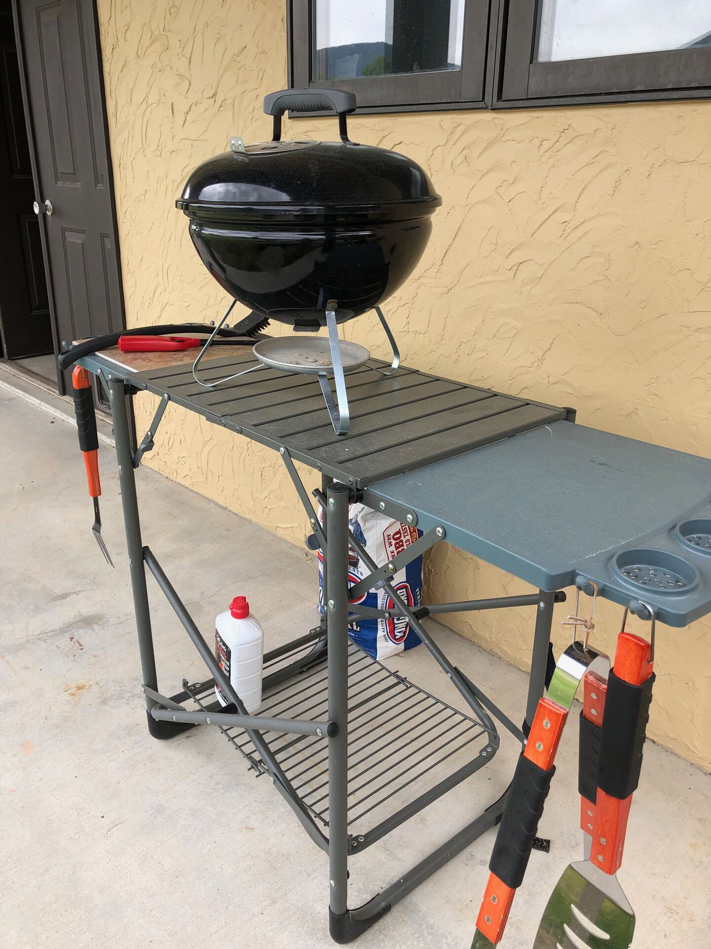 A portable barbecue grill on a metal table with a metal grill pan, a red container, and a white bottle.