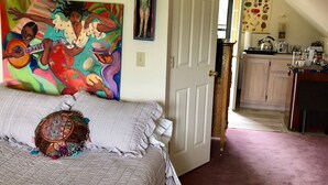 Une chambre à coucher avec un lit, un tableau coloré, une porte donnant sur la cuisine et un couloir.