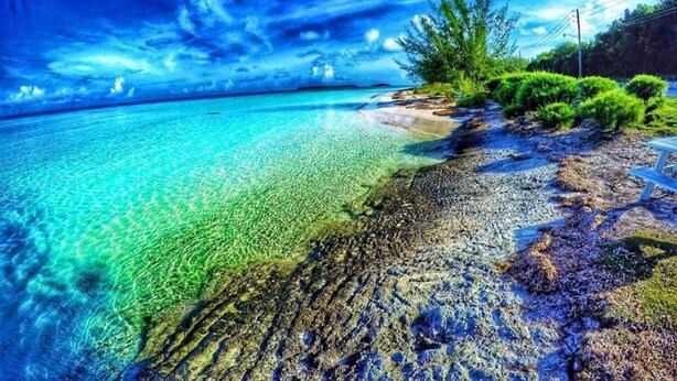 Een strand met helder turquoise water, een rotsachtige kustlijn en groene begroeiing.