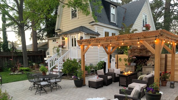 Un espace patio aménagé avec du mobilier d’extérieur, une pergola illuminée de guirlandes lumineuses et une cheminée en pierre.