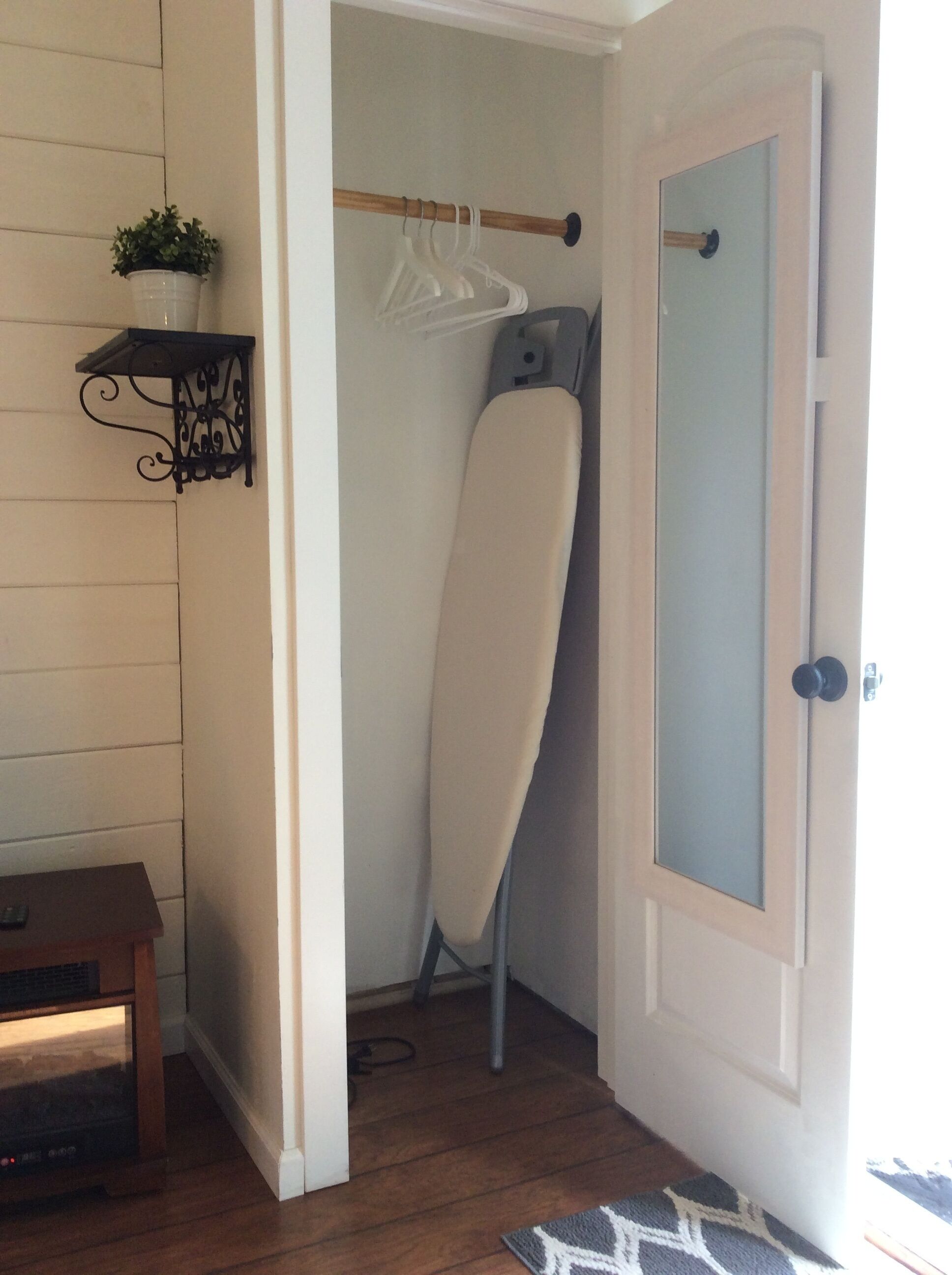 A hallway with a wooden floor, a door with a frosted glass panel, a hanging plant, a clothes rack, and a small table with a remote control.
