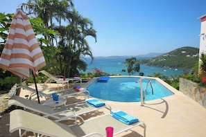 A pool area with lounge chairs, a striped umbrella, and a view of the sea.