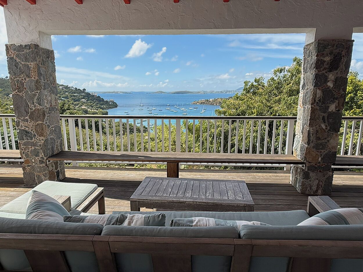 A covered outdoor seating area with stone pillars, a wooden deck, and a view of the sea and hills.