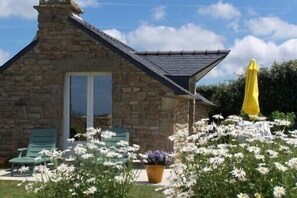 A stone building with a sloped roof, a door, and a window. There are white flowers in the foreground and a yellow umbrella in the background.