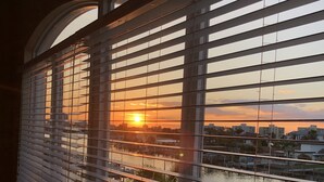 A view through window blinds of a sunset over a body of water with buildings in the background.