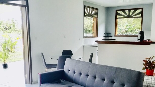 Uma sala moderna com um sofá cinzento, uma mesa de centro de vidro e uma área de jantar com balcão de cozinha.