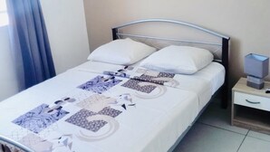Uma cama bem arrumada com colcha de padrão geométrico, uma mesinha de cabeceira com abajur e um ar condicionado na parede.