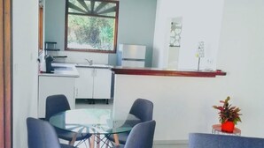 Cozinha moderna com armários brancos, área de jantar com mesa de vidro e quatro cadeiras, e um sofá azul com detalhe vermelho.