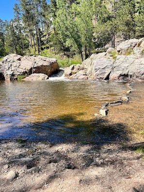 Entorno natural al aire libre con un cuerpo de agua cristalina, terreno rocoso y árboles al fondo.