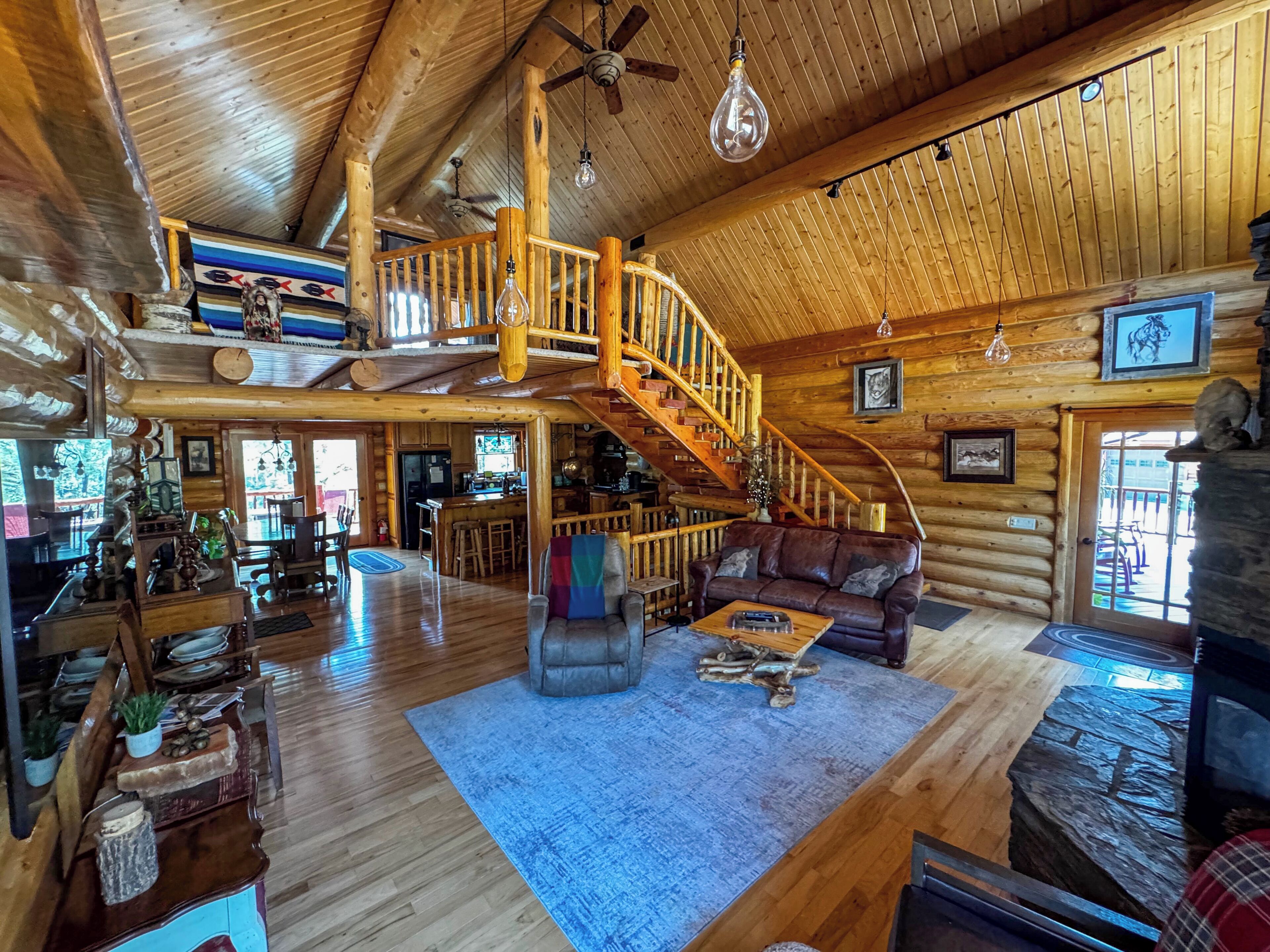A spacious log cabin interior with a wooden staircase, a cozy seating area, and a kitchen visible through an open door.
