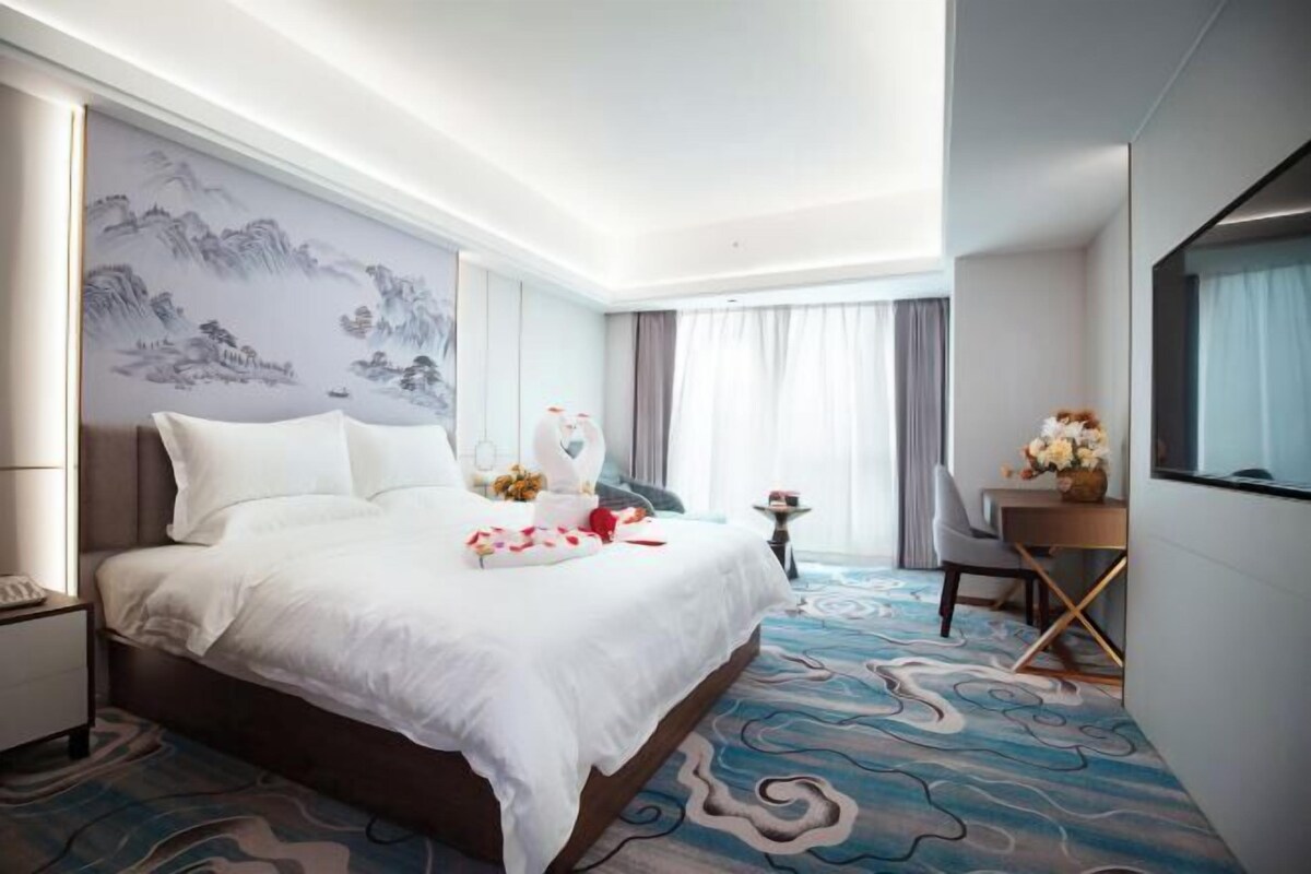A hotel room with a large bed, a desk, and a chair. There is a painting on the wall and a window with curtains.