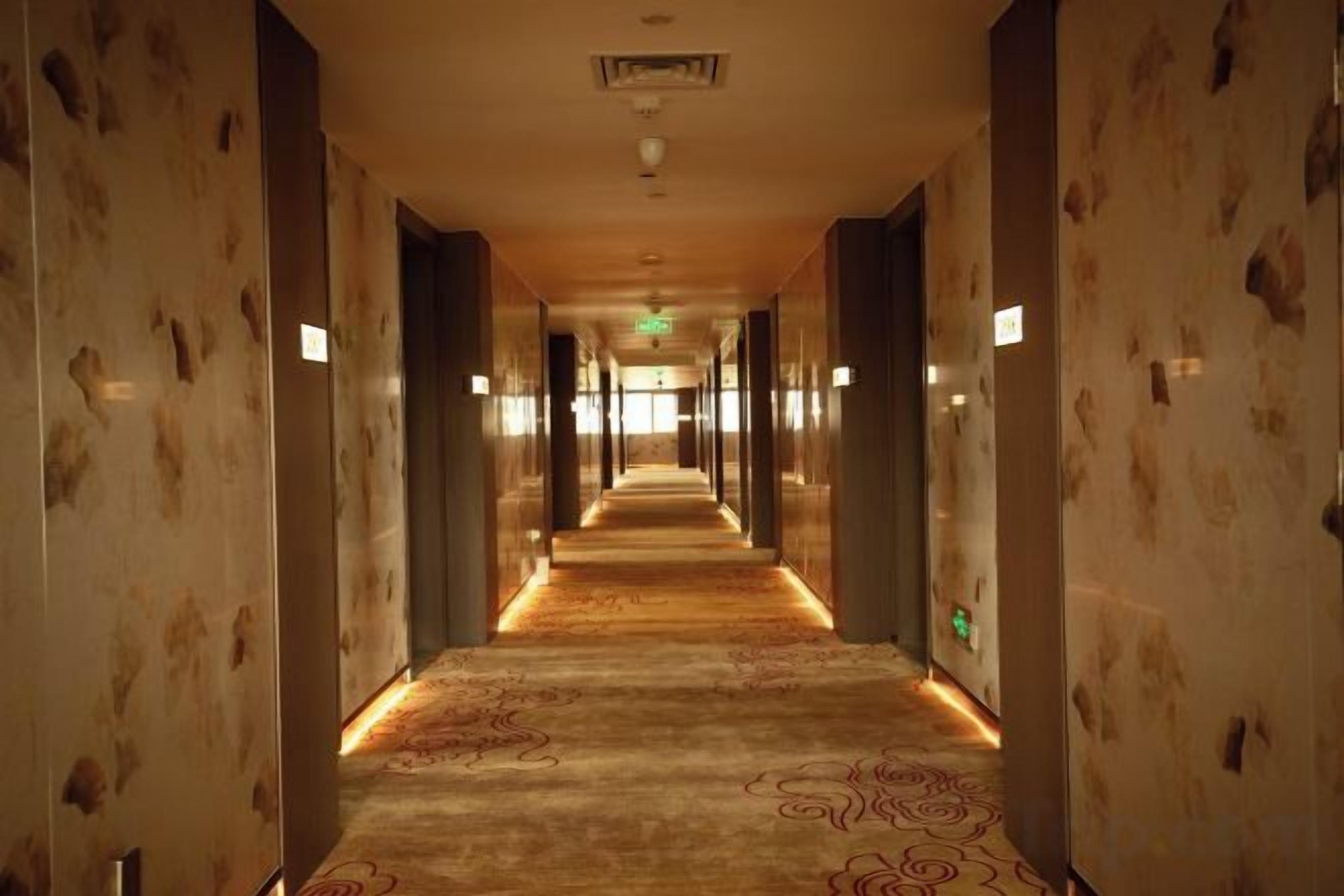 A hotel hallway with carpeted floors, doors on both sides, and a long corridor leading to a bright area at the end.