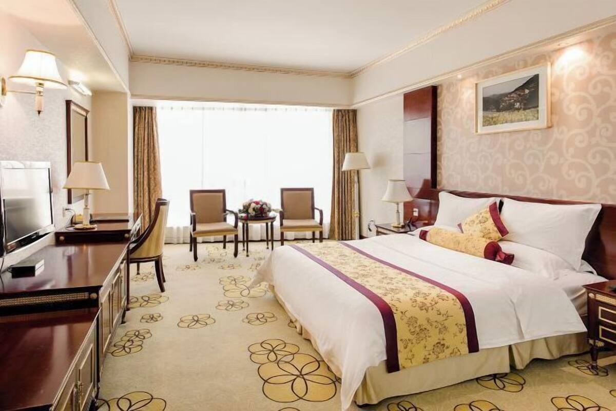 Une chambre d’hôtel comprenant un lit, un bureau, une télévision, deux chaises, une petite table et une fenêtre avec des rideaux.
