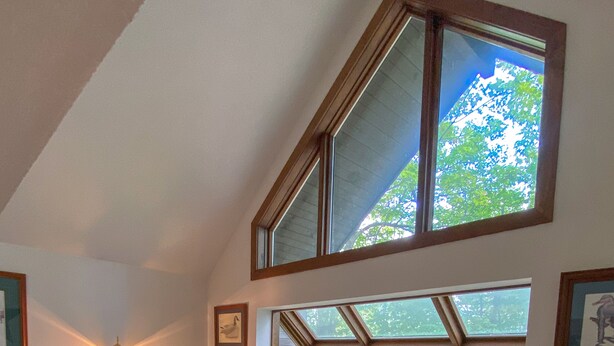 Ein geräumiges Wohnzimmer mit hoher Decke, großen Fenstern mit Blick ins Freie, einer braunen Ledersitzgruppe und einem Holztisch.