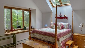 Ein Schlafzimmer mit Himmelbett, Dachfenster, einer Holzkommode, einem Nachttisch und einem Fenster mit Blick auf Bäume.
