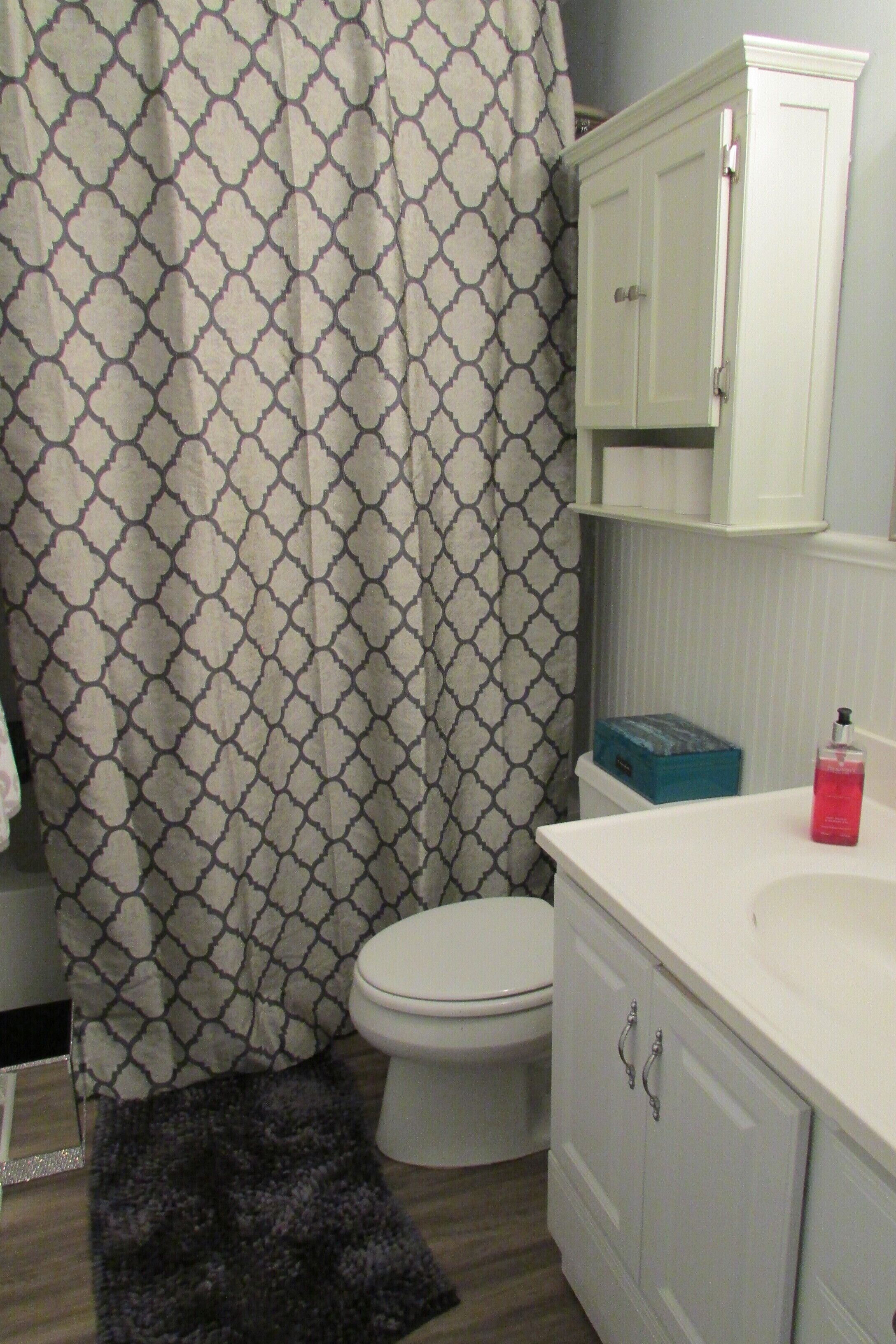 Un baño con cortina de ducha estampada, un inodoro blanco, un lavabo blanco con un jabonera azul y un armario blanco con tiradores plateados.