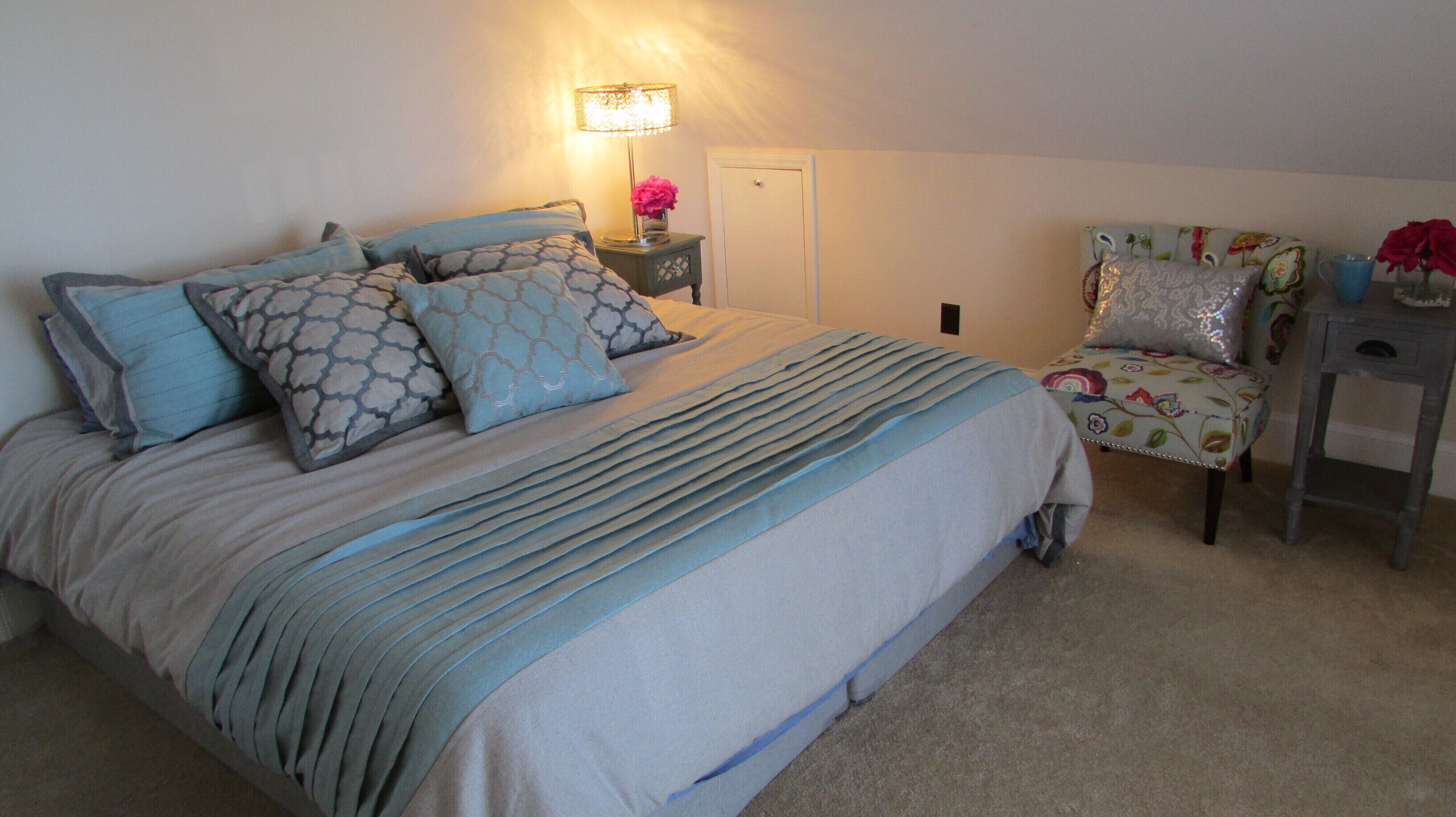 A neatly made bed with a striped bedspread and multiple pillows, a bedside table with a lamp and a vase of flowers, a floral chair, and a small table with a vase of red roses.