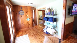 A hallway with wooden walls and floors, a mirror, a shelf with folded clothes, and a television screen.