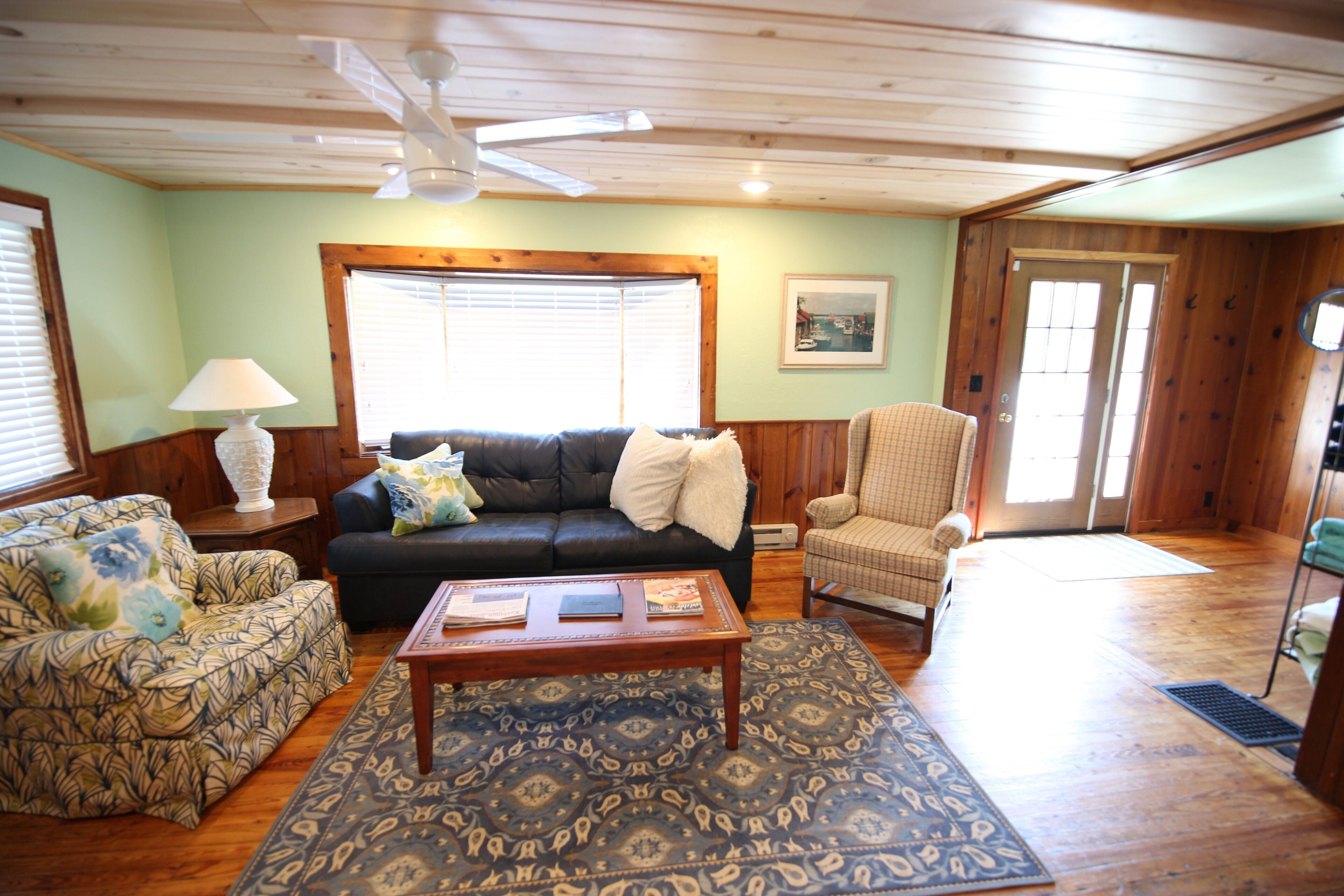A living room with a sofa, armchair, and coffee table, wooden floors, and a ceiling fan.