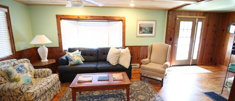 A living room with a sofa, armchair, and coffee table, wooden floors, and a ceiling fan.