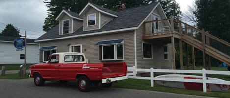 Une maison à deux étages avec une terrasse en bois et un camion rouge garé devant.