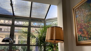 Una habitación iluminada por el sol con techo de cristal, una mesa con sillas, una lámpara, una planta en maceta y un cuadro en la pared.