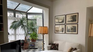 Una sala de estar con un sofá, un reposapiés, un televisor, plantas en macetas y cuadros en la pared.