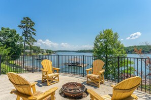 Une terrasse dotée de chaises Adirondack et d’un foyer extérieur, offrant une vue sur un lac où l’on peut apercevoir des bateaux et des quais.