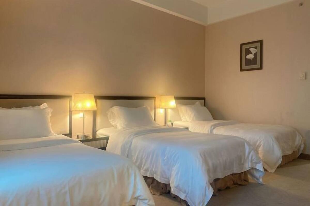 Een hotelkamer met drie bedden, elk voorzien van witte beddengoed en hoofdeinden. Er staan nachtlampjes op elk nachtkastje.