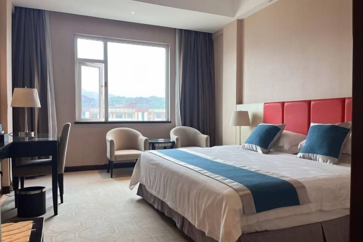 Hotelkamer met een bed, twee stoelen, een bureau, een lamp en uitzicht op een gebouw door een raam.