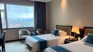 Ein Hotelzimmer mit zwei Betten, einem Schreibtisch, einem Stuhl, einer Lampe und Blick auf eine Stadtlandschaft.